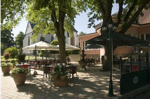 Outdoor patio with tables and chairs shaded by large trees and umbrellas, surrounded by potted plants. A pink building and a church with a tall roof are visible in the background on a sunny day.