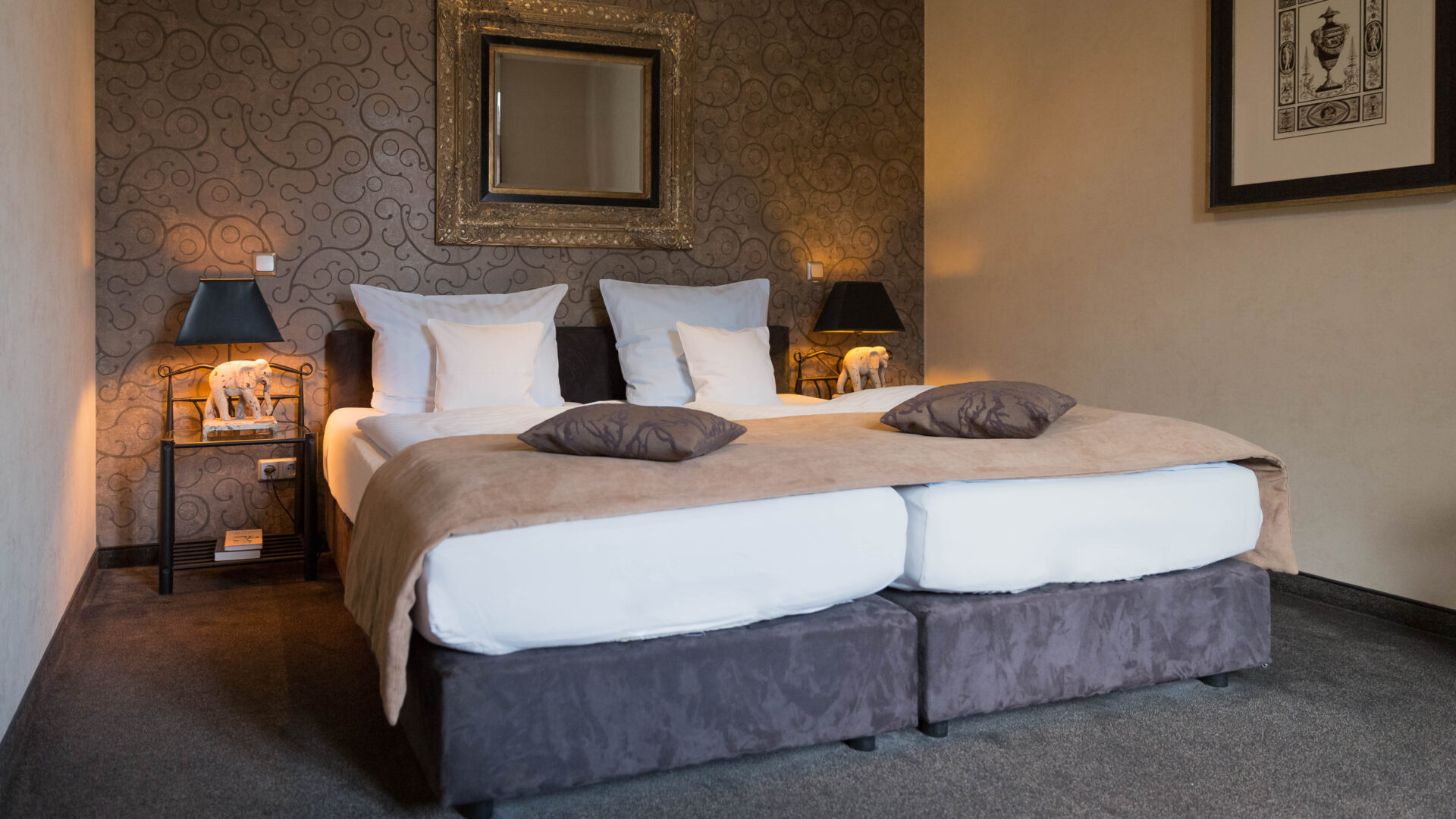 A neatly made hotel bed with white linens and tan blankets, two brown pillows, bedside tables with lamps, patterned accent wall, framed mirror, and framed artwork on the adjacent wall.