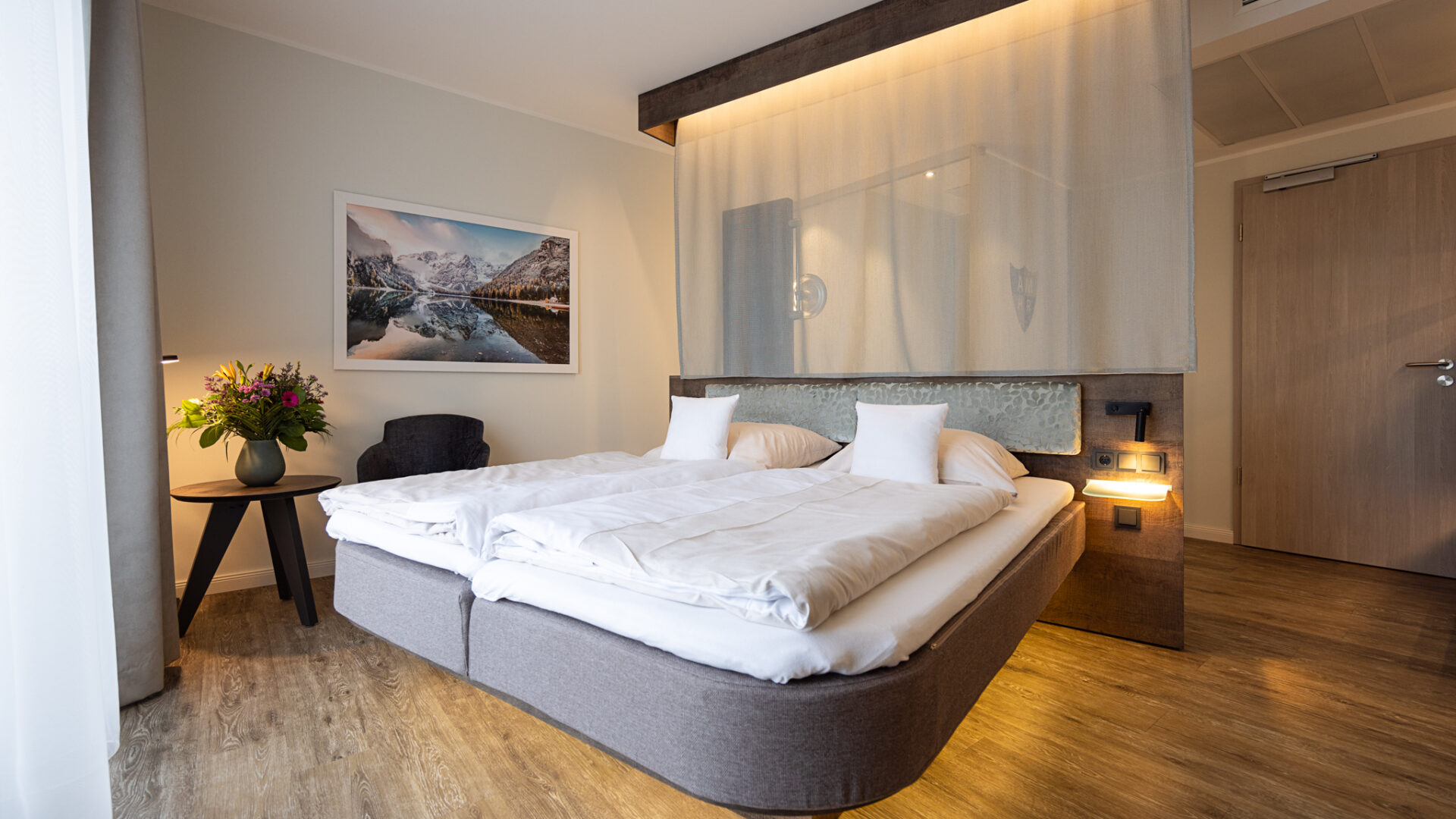 A modern hotel room with a large bed, white bedding, two pillows, wooden flooring, a side table with a bouquet of flowers, a gray armchair, and a wall-mounted landscape photograph above the bed.