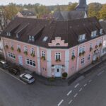 Aerial view of a pink, three-story corner building with a dark roof and flower boxes under the windows, located at a curved intersection with parked cars and surrounded by trees and other houses.
