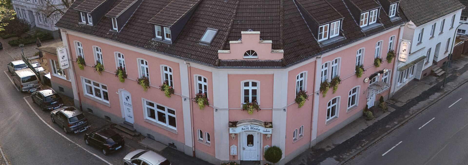 Aerial view of a pink, three-story corner building with a dark roof and flower boxes under the windows, located at a curved intersection with parked cars and surrounded by trees and other houses.