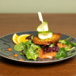 A gourmet salad with greens, smoked salmon, a crisp potato cake, and cucumber slices on a gray plate, garnished with orange slices and sauce dots, presented on a wooden table.