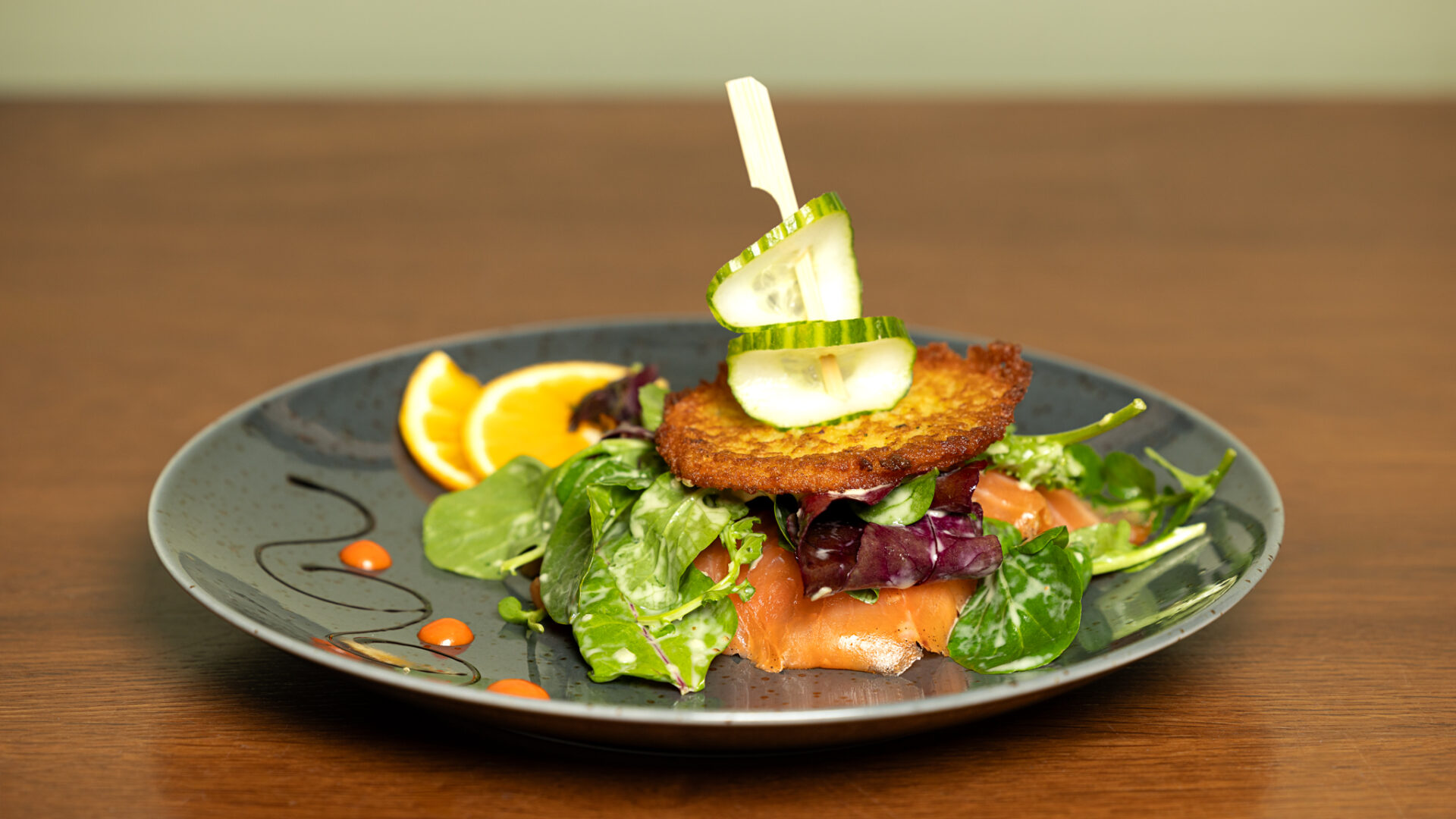 A gourmet salad with greens, smoked salmon, a crisp potato cake, and cucumber slices on a gray plate, garnished with orange slices and sauce dots, presented on a wooden table.