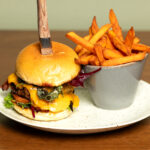 A cheeseburger with lettuce, tomato, and cheddar cheese sits on a plate, with a knife stuck into the bun. Next to it is a bowl filled with sweet potato fries. The plate is on a wooden table.
