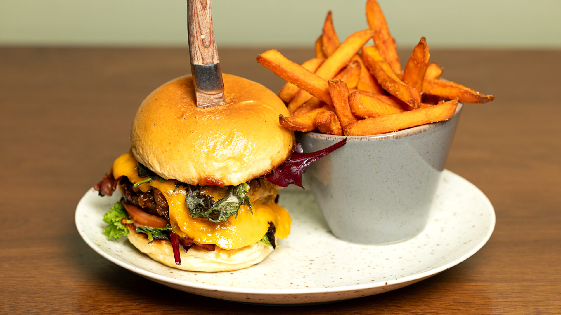 A cheeseburger with lettuce, tomato, and cheddar cheese sits on a plate, with a knife stuck into the bun. Next to it is a bowl filled with sweet potato fries. The plate is on a wooden table.