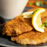 Two breaded and fried schnitzels garnished with lemon slices and herbs, served on a dark plate with a small black dish of red sauce and a blurred bowl in the background.