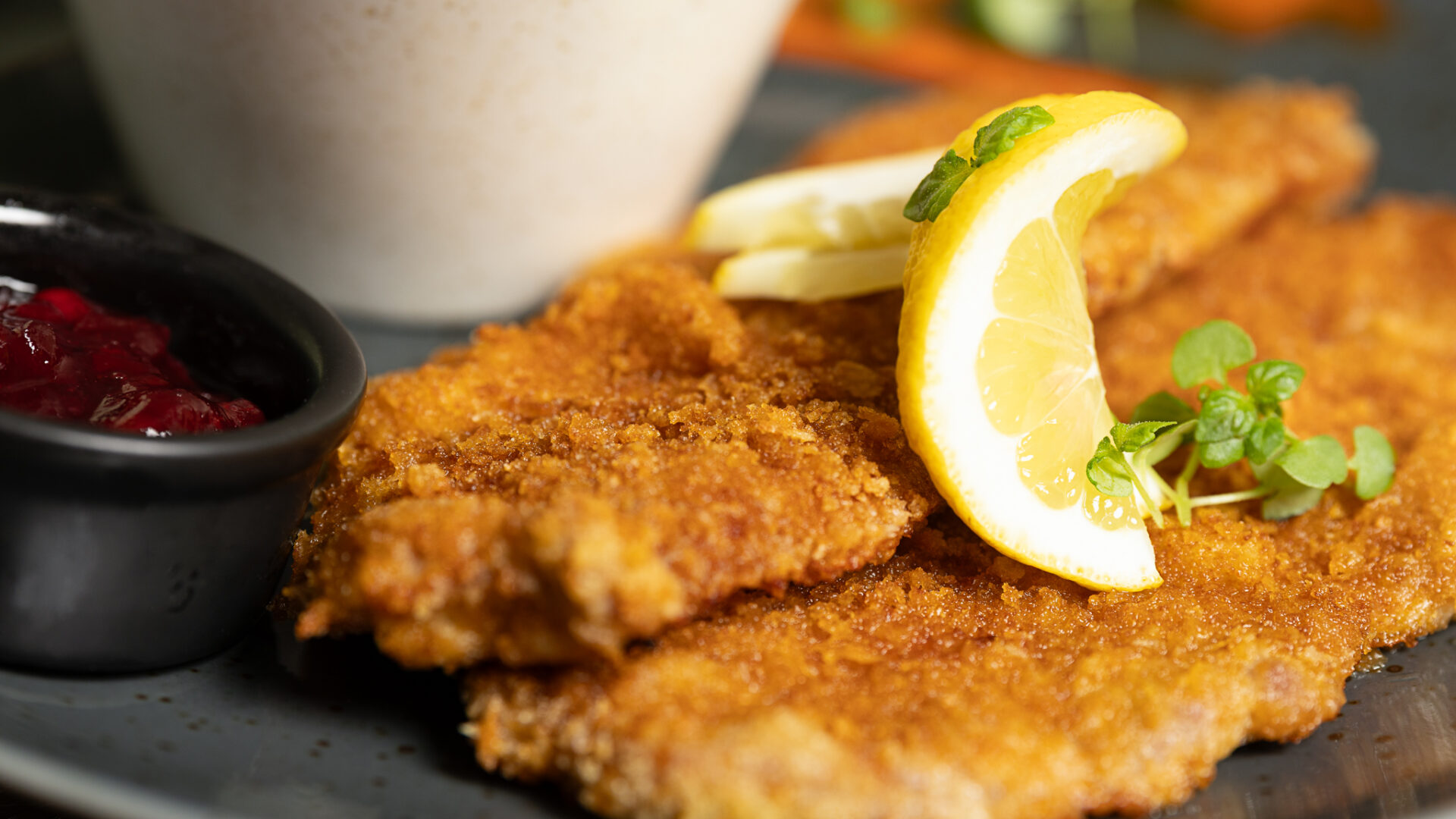 Two breaded and fried schnitzels garnished with lemon slices and herbs, served on a dark plate with a small black dish of red sauce and a blurred bowl in the background.