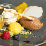 A gourmet dessert plate with a scoop of vanilla ice cream in a glass jar, a chocolate mousse quenelle, yellow cream, a pastry stick, and garnishes of raspberry, blueberry, physalis, and sauce on a gray plate.