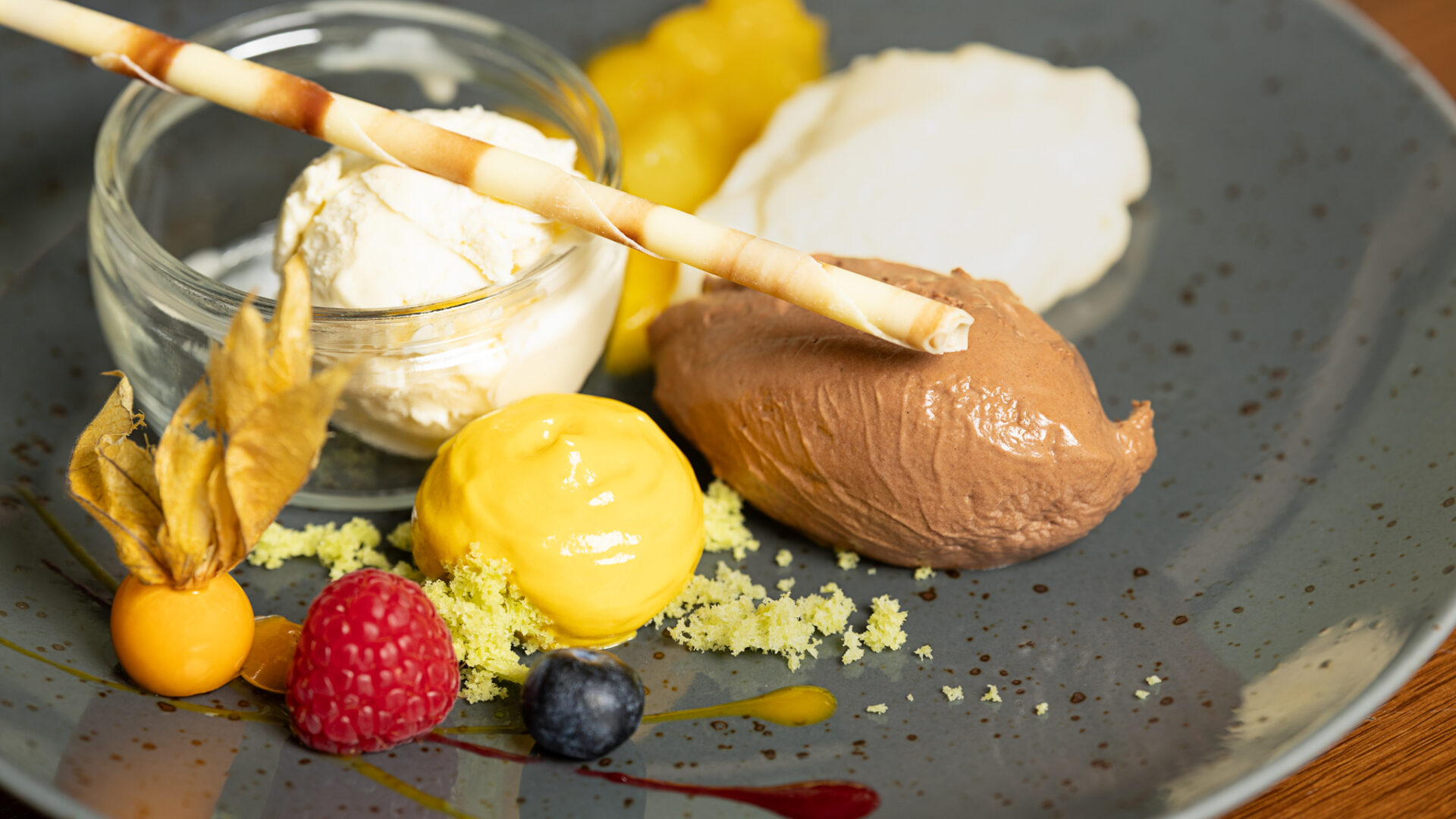 A gourmet dessert plate with a scoop of vanilla ice cream in a glass jar, a chocolate mousse quenelle, yellow cream, a pastry stick, and garnishes of raspberry, blueberry, physalis, and sauce on a gray plate.