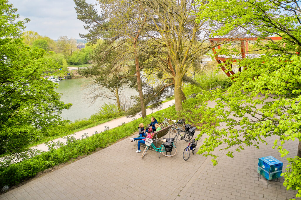 A group of people sits on benches next to bicycles under leafy trees near a river, with a paved path and green foliage surrounding them on a bright day.