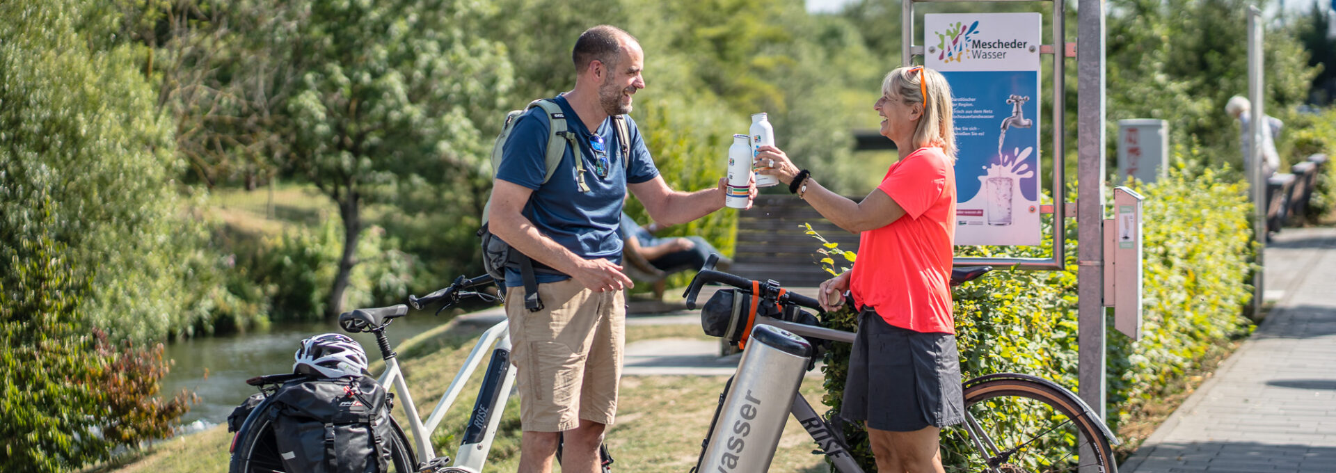Two cyclists, a man and a woman, refill their water bottles at an outdoor drinking water station near a bike path. Both are smiling, standing beside their bikes, with greenery and informational signs in the background.