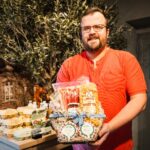 A man in a red shirt smiles while holding a gift basket filled with assorted food items, standing indoors next to a display of eggs and other products with a leafy plant in the background.