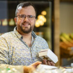 A bearded man with glasses, wearing a patterned shirt, smiles while holding a small open box in a shop with fresh produce and jars visible in the background.
