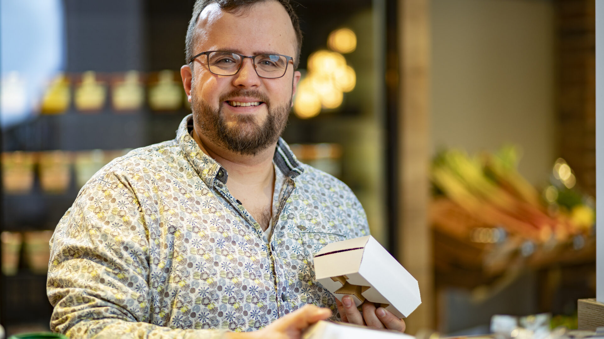 A bearded man with glasses, wearing a patterned shirt, smiles while holding a small open box in a shop with fresh produce and jars visible in the background.