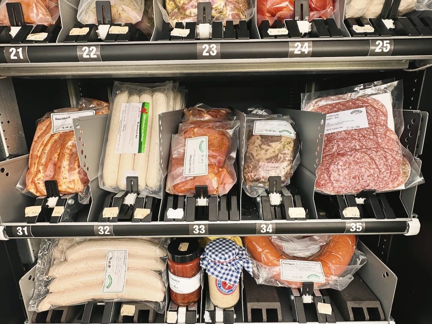 A vending machine filled with various packaged meats, sausages, and jars, organized on metal shelves labeled with numbers, offering a selection of deli items in a self-serve display.