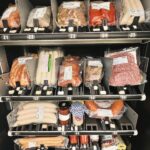 A vending machine filled with various packaged meats, sausages, and jars, organized on metal shelves labeled with numbers, offering a selection of deli items in a self-serve display.