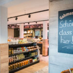 A welcoming local shop with shelves of assorted food products and a chalkboard sign reading “Schön dass Du hier bist!” (translation: “Nice that you’re here!”) by the entrance.