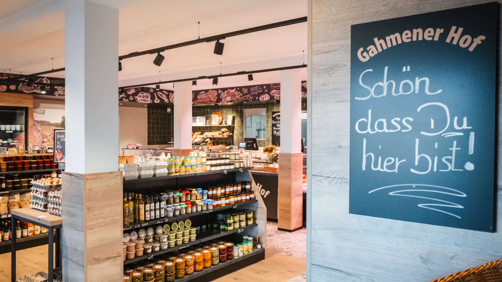 A welcoming local shop with shelves of assorted food products and a chalkboard sign reading “Schön dass Du hier bist!” (translation: “Nice that you’re here!”) by the entrance.