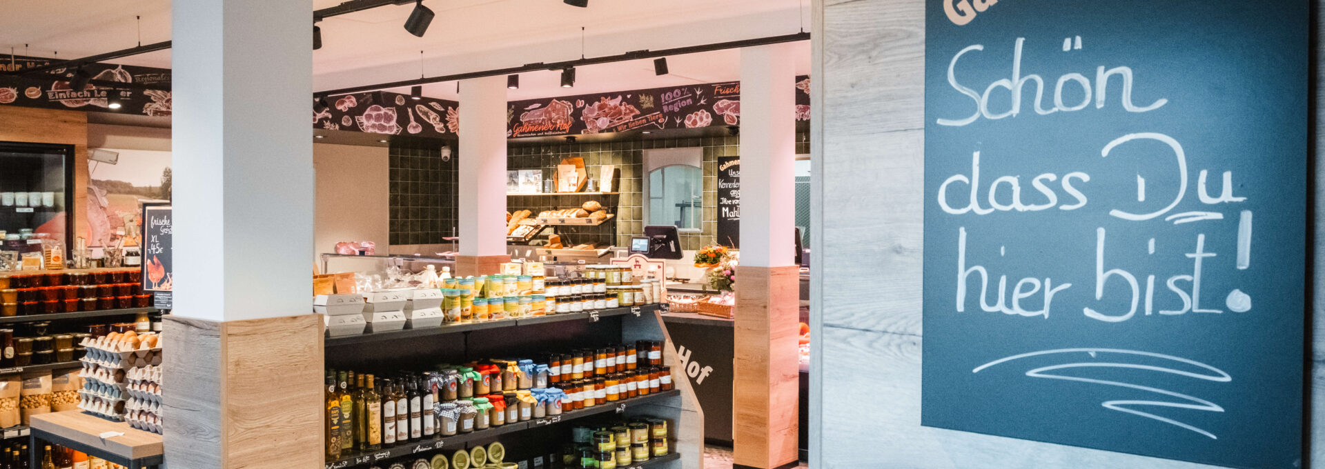 A welcoming local shop with shelves of assorted food products and a chalkboard sign reading “Schön dass Du hier bist!” (translation: “Nice that you’re here!”) by the entrance.