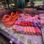 A butcher shop display case filled with various sausages and deli meats, neatly arranged in rows with German labels and prices visible in front of each type. Baskets and chalkboard menus are seen in the background.