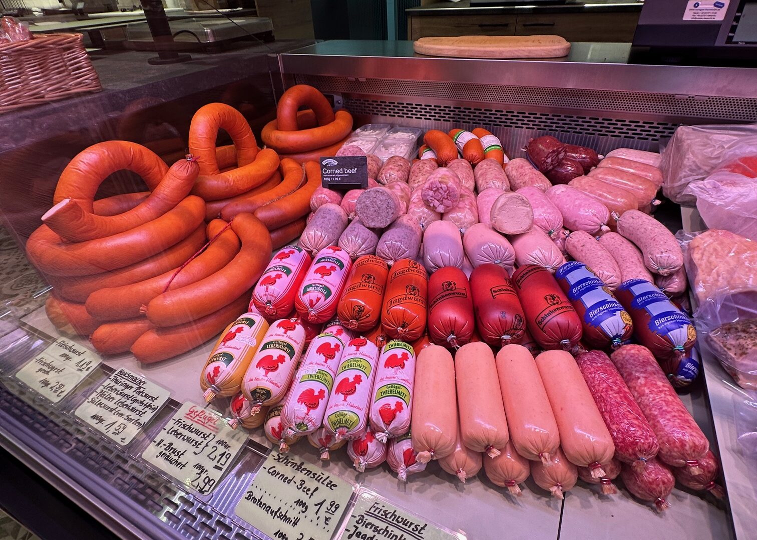 A butcher shop display case filled with various sausages and deli meats, neatly arranged in rows with German labels and prices visible in front of each type. Baskets and chalkboard menus are seen in the background.