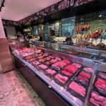 A modern butcher shop with a long glass display case filled with various cuts of raw meat. Behind the counter, sausages and cured meats hang on hooks, and chalkboard-style drawings decorate the walls.