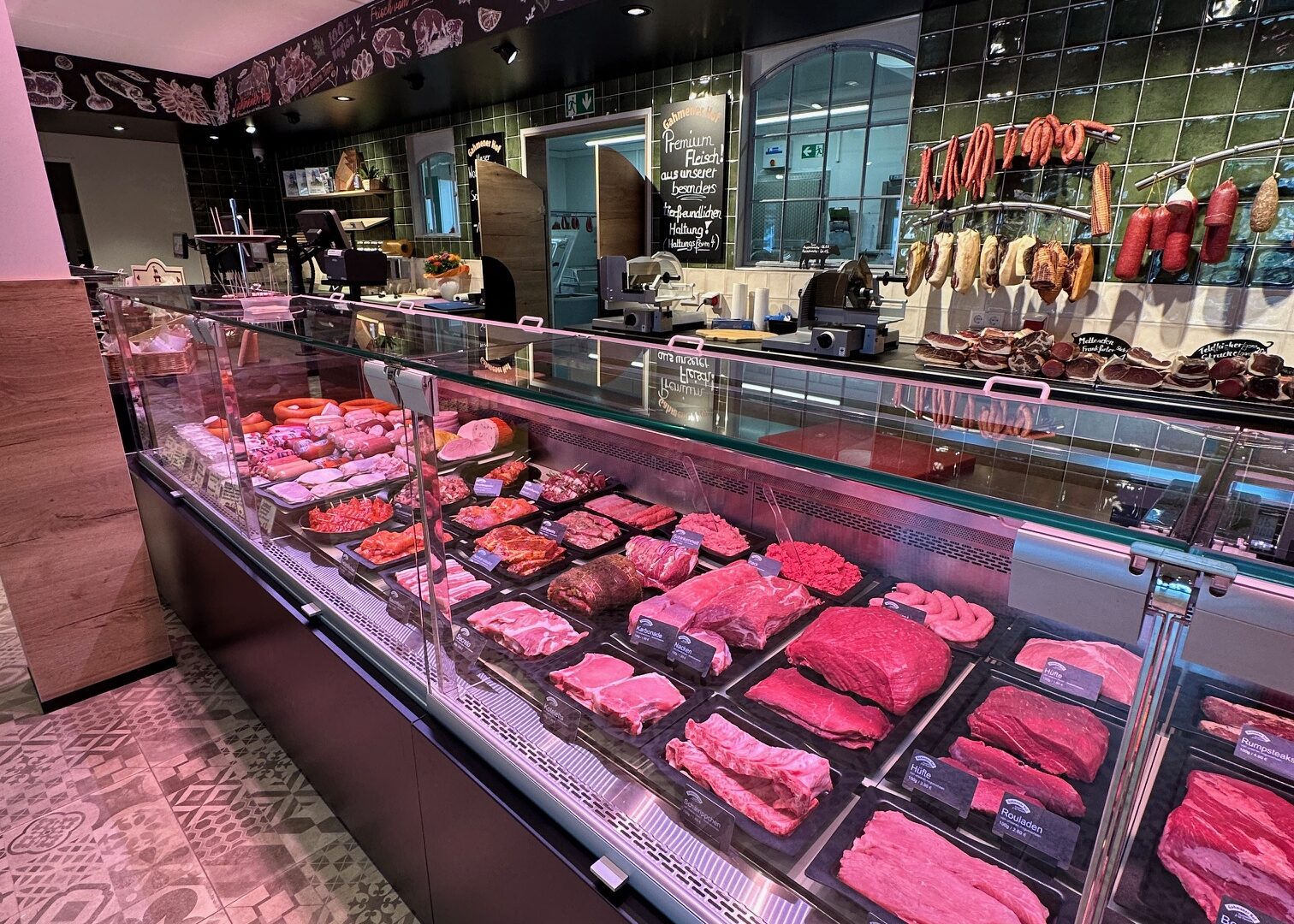 A modern butcher shop with a long glass display case filled with various cuts of raw meat. Behind the counter, sausages and cured meats hang on hooks, and chalkboard-style drawings decorate the walls.
