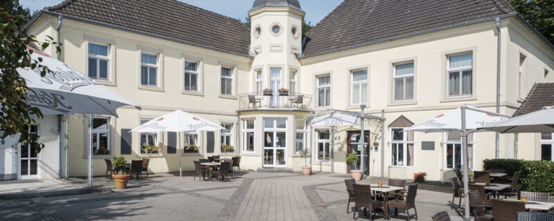 A large, pale yellow building with a central tower and tiled roof surrounds an open courtyard with outdoor tables, chairs, and umbrellas. The scene is bright and inviting, with clear skies above.