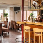 A cozy, modern cafe with brown upholstered chairs, wooden tables, a bar with high stools, potted plants, and shelves stocked with bottles and glasses. Large windows let in natural light.