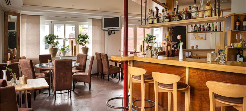 A cozy, modern cafe with brown upholstered chairs, wooden tables, a bar with high stools, potted plants, and shelves stocked with bottles and glasses. Large windows let in natural light.