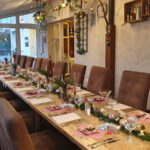 A long dining table set for a festive meal with brown chairs, decorated with candles, greenery, and ornaments. Place settings include napkins, menus, and glasses. Holiday decorations hang from the ceiling and walls.