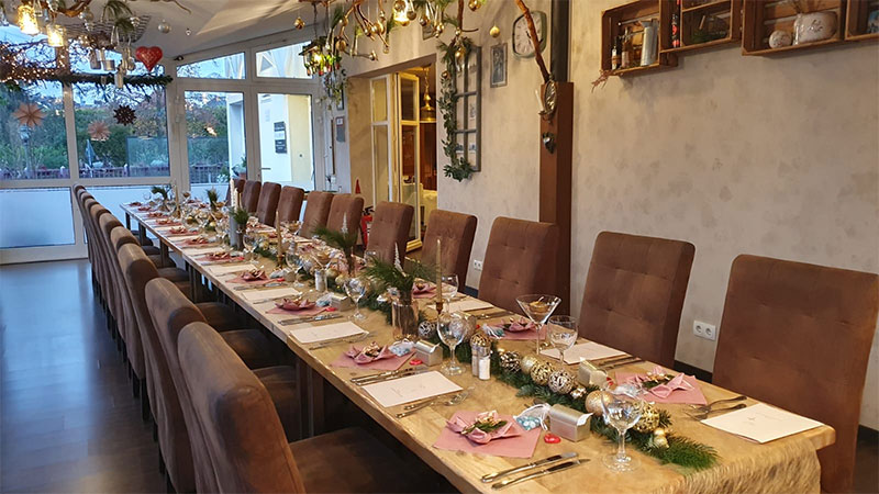 A long dining table set for a festive meal with brown chairs, decorated with candles, greenery, and ornaments. Place settings include napkins, menus, and glasses. Holiday decorations hang from the ceiling and walls.