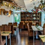 A cozy restaurant with brown chairs, wooden tables, and festive holiday decorations including hanging branches, ornaments, and greenery. Large windows line one side, letting in natural light. The atmosphere is warm and inviting.