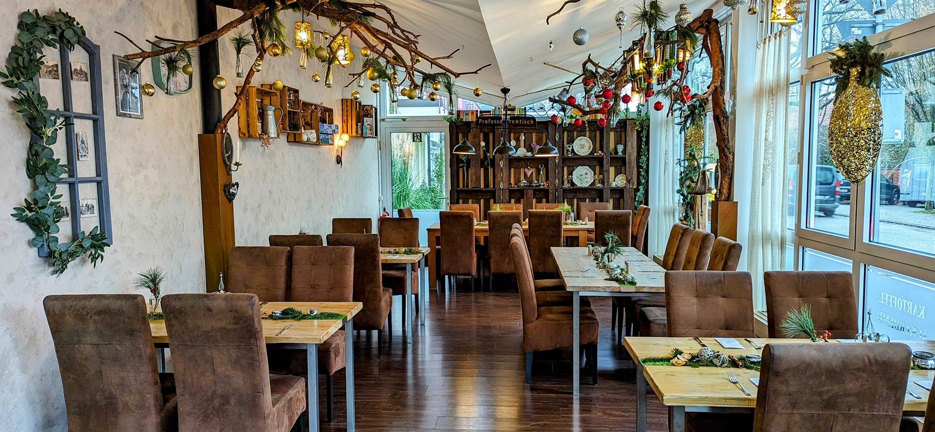 A cozy restaurant with brown chairs, wooden tables, and festive holiday decorations including hanging branches, ornaments, and greenery. Large windows line one side, letting in natural light. The atmosphere is warm and inviting.