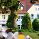 A charming yellow hotel and restaurant with red roof tiles, surrounded by lush green trees and flowering plants in the foreground, on a sunny day.