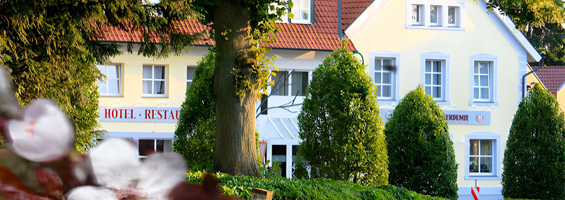 A charming yellow hotel and restaurant with red roof tiles, surrounded by lush green trees and flowering plants in the foreground, on a sunny day.