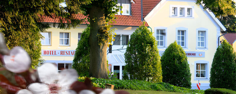 A charming yellow hotel and restaurant with red roof tiles, surrounded by lush green trees and flowering plants in the foreground, on a sunny day.