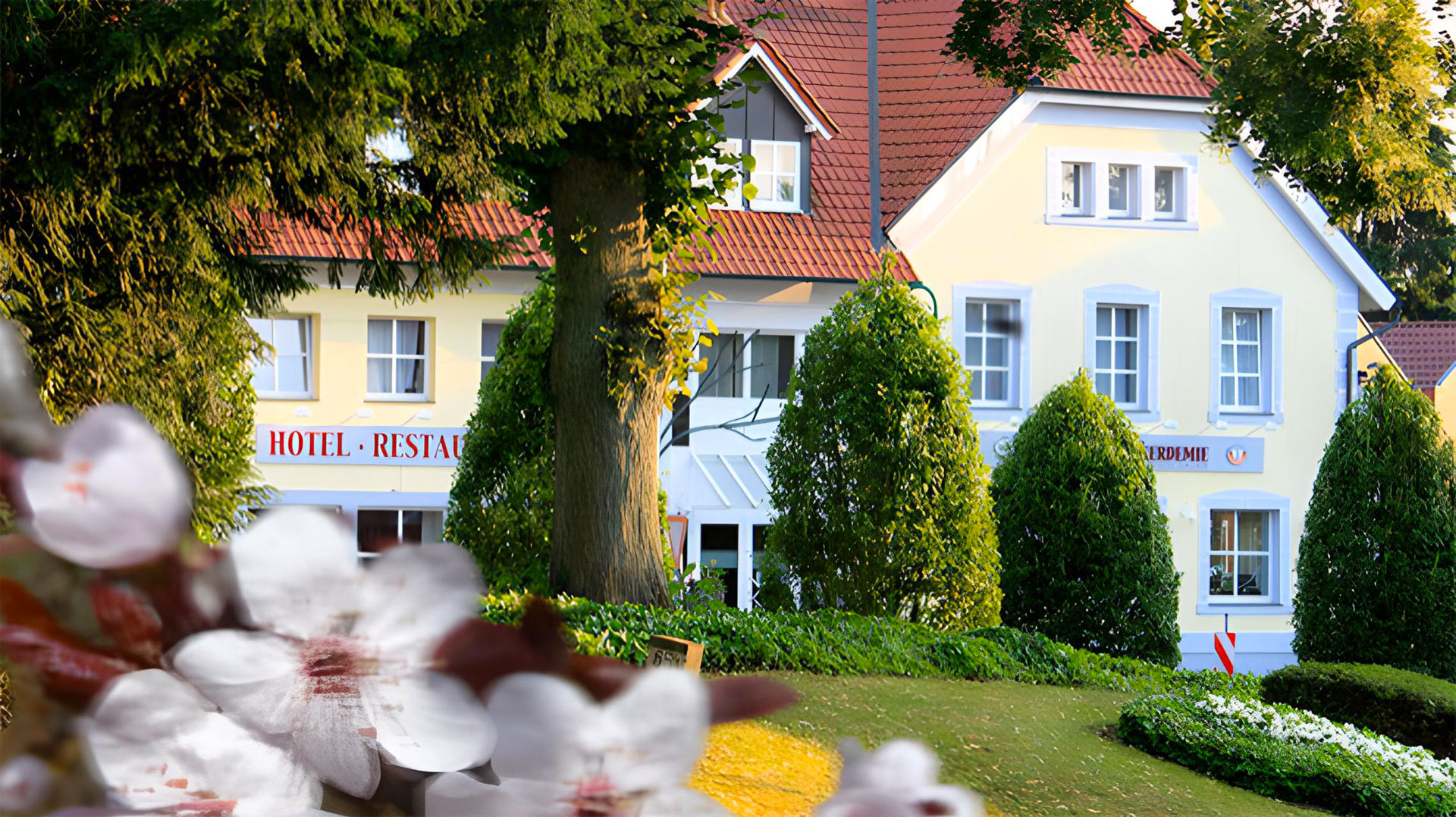 A charming yellow hotel and restaurant with red roof tiles, surrounded by lush green trees and flowering plants in the foreground, on a sunny day.