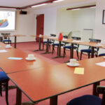 A conference room with U-shaped tables, blue chairs, cups and saucers, notepads, and pens. A projector displays charts and graphs on a screen at the front of the room. The room is brightly lit.