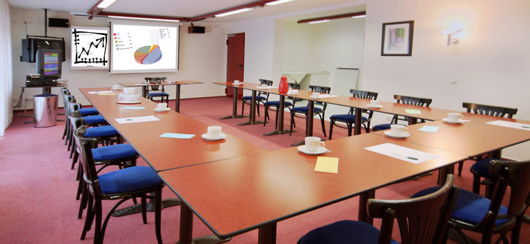 A conference room with U-shaped tables, blue chairs, cups and saucers, notepads, and pens. A projector displays charts and graphs on a screen at the front of the room. The room is brightly lit.
