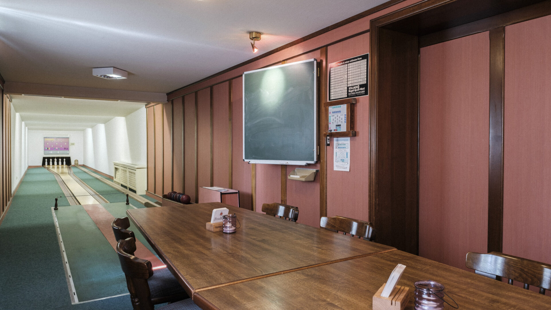 A long wooden table with chairs is set in a room beside a small bowling lane. A chalkboard hangs on the wall above the table, and the space has pink walls with wooden trim and a ceiling light.