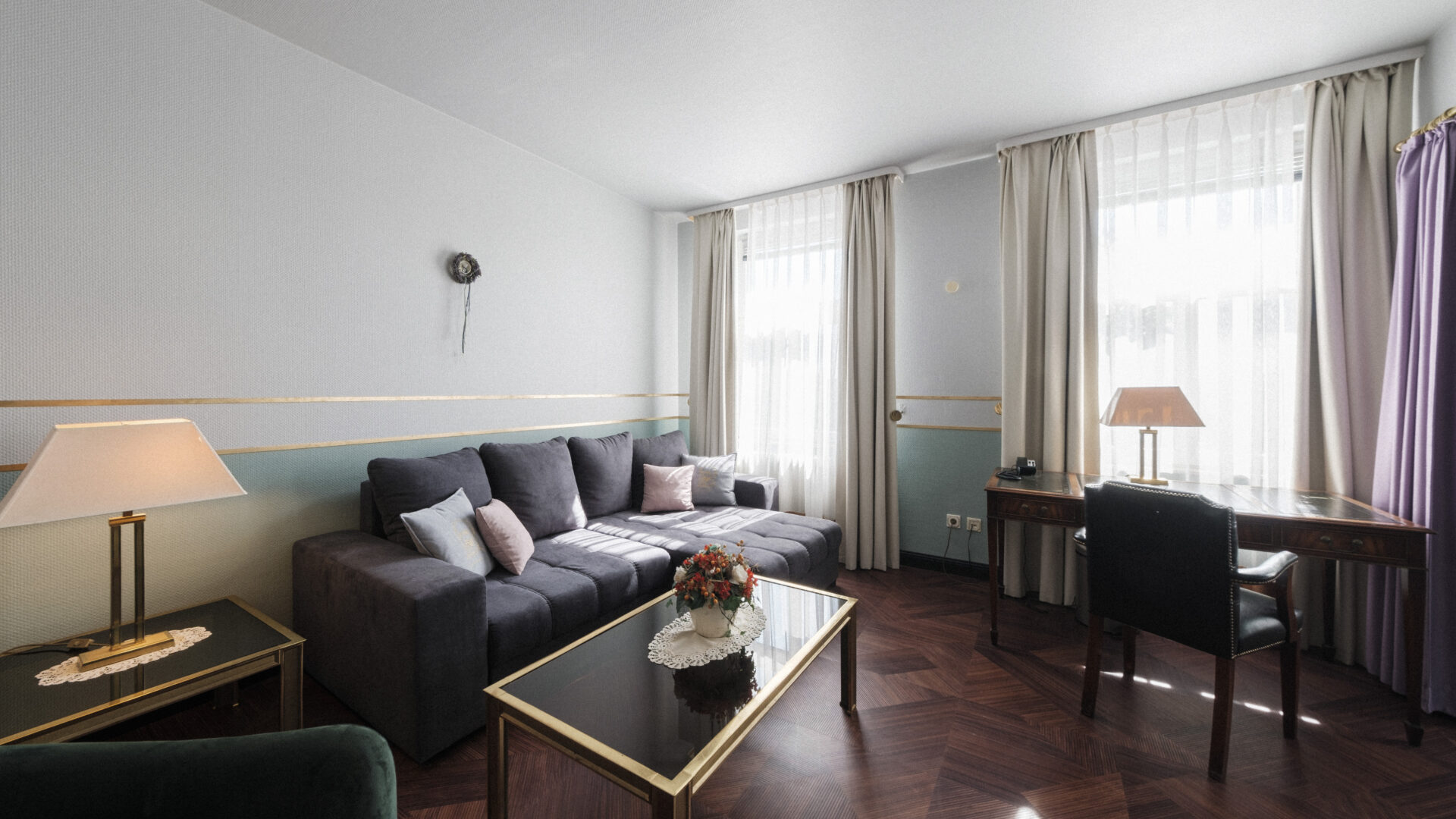 A bright living room with a dark gray sofa, glass coffee table, desk and chair by two large windows with sheer curtains, and a table lamp. The floor is dark wood with a vase of flowers on the coffee table.
