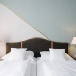 Two single beds with white bedding are placed side by side, each with two white pillows. The beds have a shared dark headboard. Two wall lamps are mounted above wooden nightstands on either side. The wall is painted two tones.