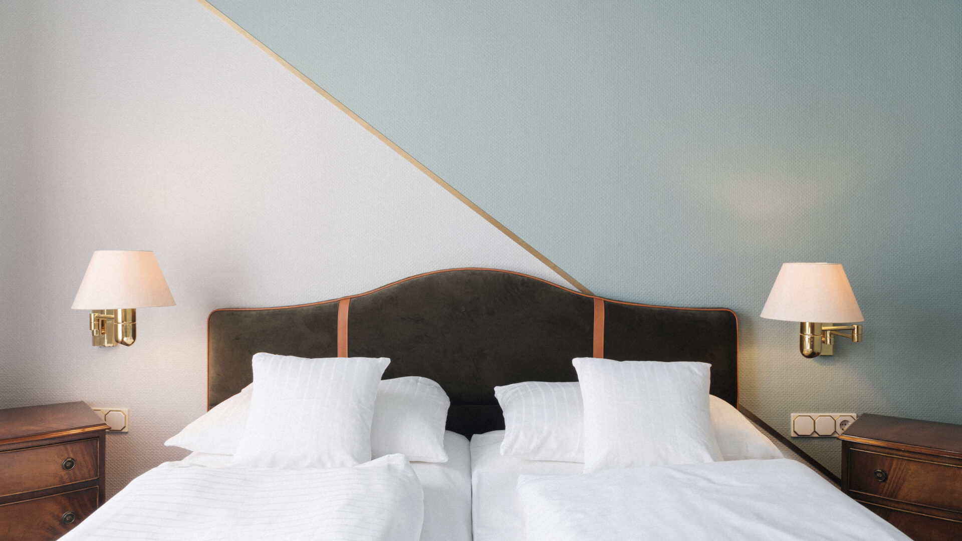 Two single beds with white bedding are placed side by side, each with two white pillows. The beds have a shared dark headboard. Two wall lamps are mounted above wooden nightstands on either side. The wall is painted two tones.