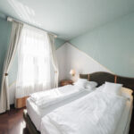 A bright, modern hotel room with two single beds pushed together, white bedding, light blue walls, wooden nightstands, wall-mounted lamps, and large window with sheer curtains.