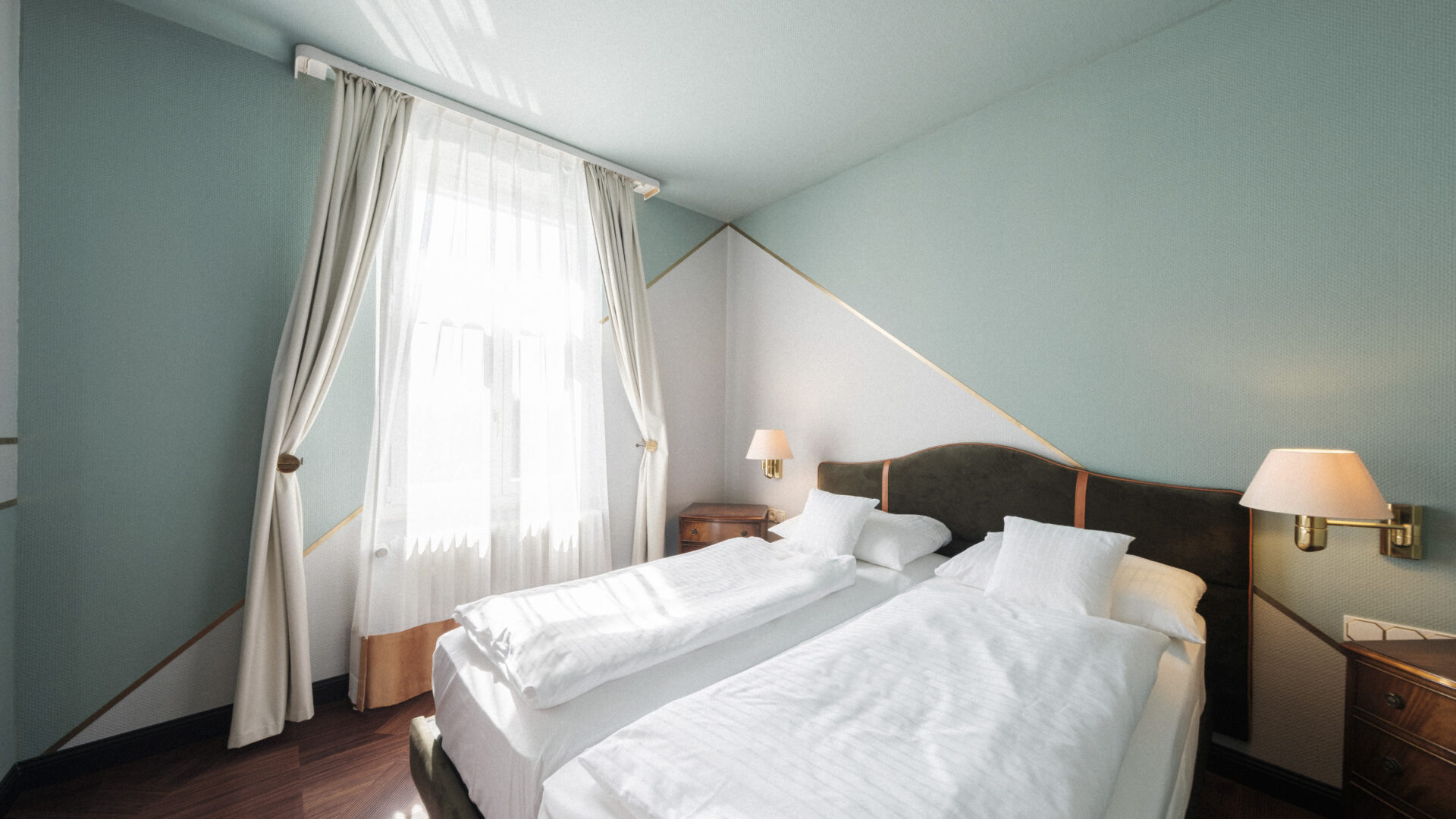 A bright, modern hotel room with two single beds pushed together, white bedding, light blue walls, wooden nightstands, wall-mounted lamps, and large window with sheer curtains.