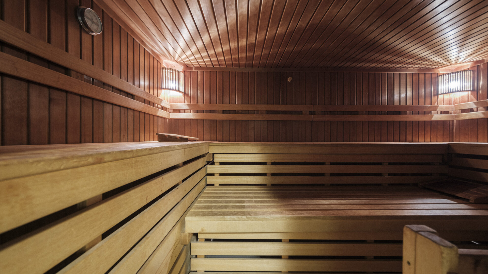 A spacious wooden sauna with tiered benches, warm lighting, and a clock on the wall, creating a relaxing and cozy atmosphere.