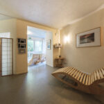 A modern, minimalist room with light walls, a wooden lounge chair, a folding screen, a framed picture, and a small table; glass doors at the back open to a sunlit area with two wooden rocking chairs.