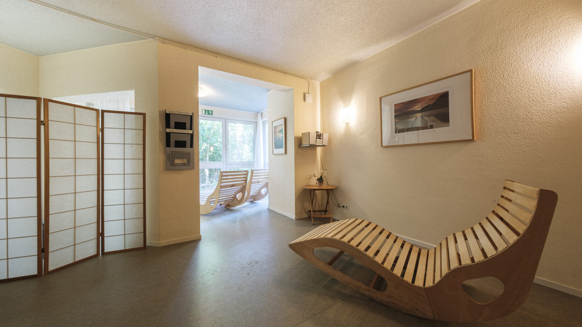 A modern, minimalist room with light walls, a wooden lounge chair, a folding screen, a framed picture, and a small table; glass doors at the back open to a sunlit area with two wooden rocking chairs.