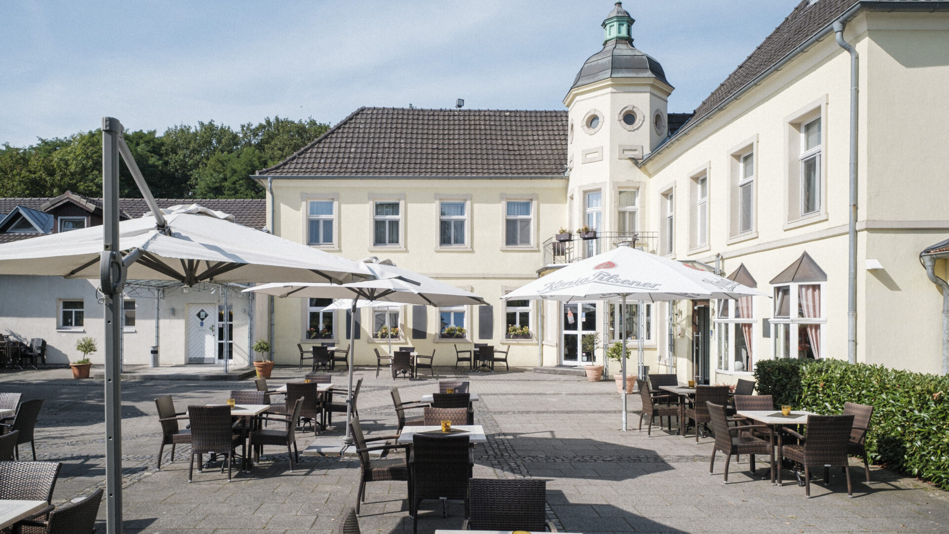 Outdoor seating area with wicker chairs and tables, some under large white umbrellas, in front of a cream-colored building with a tower and green roof on a sunny day.